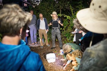 Een bodemdierengids met safarihoed neemt een groep mensen in het donker mee het Arboretum in. De mensen luisteren en sommigen zijn al bodemdieren aan het zoeken